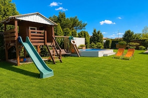 A wooden playhouse with a slide, a small pool, and two lounge chairs in a grassy backyard. - Cottages for 4 People With a Swimming Pool (Kamien Pomorski)
