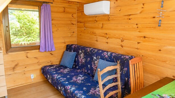 A wooden cabin room with a floral sofa, a window with a purple curtain, and an air conditioning unit. - Cottages for 4 People With a Swimming Pool (Kamien Pomorski)