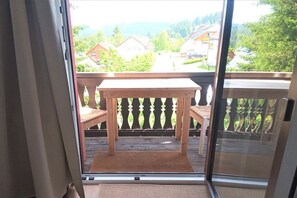 Un balcón con mesa y sillas de madera, vistas a casas y follaje, y una puerta que da al interior.