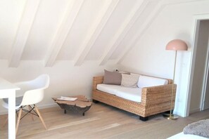 Una habitación estilo loft con techo blanco, sofá de mimbre con cojines, una silla blanca, una pequeña mesa de centro de madera y una lámpara de pie alta.