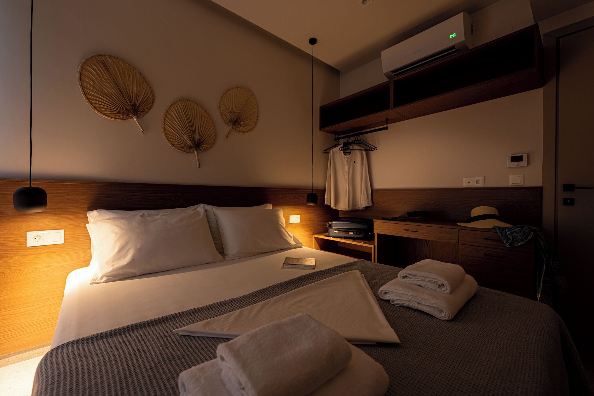 Habitación de hotel con cama, mesita de noche, ventilador de pared y estante con sombrero y un libro.