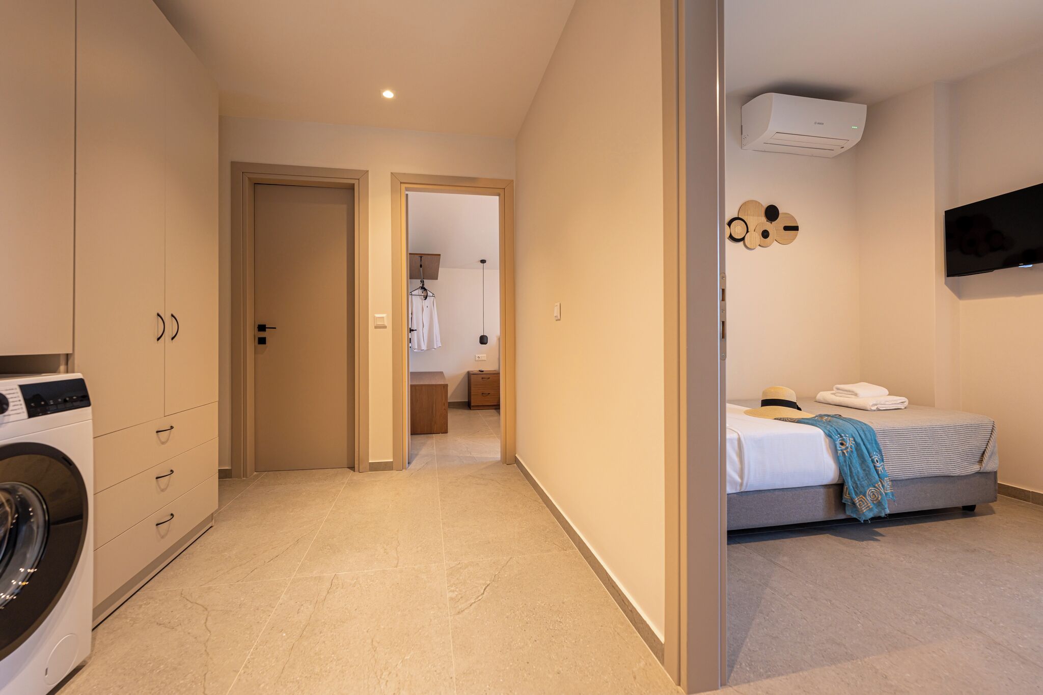 A hotel room with a bed, a TV, a washing machine, and a hallway leading to another room.