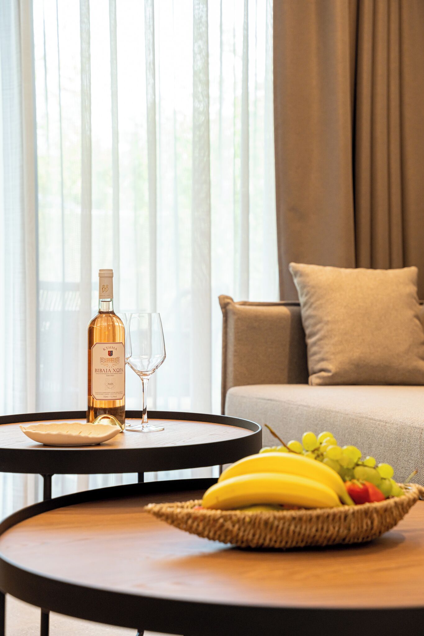 A bottle of Bibaia XO wine, a glass, and a fruit basket on a round table in a room with a sofa and curtains.