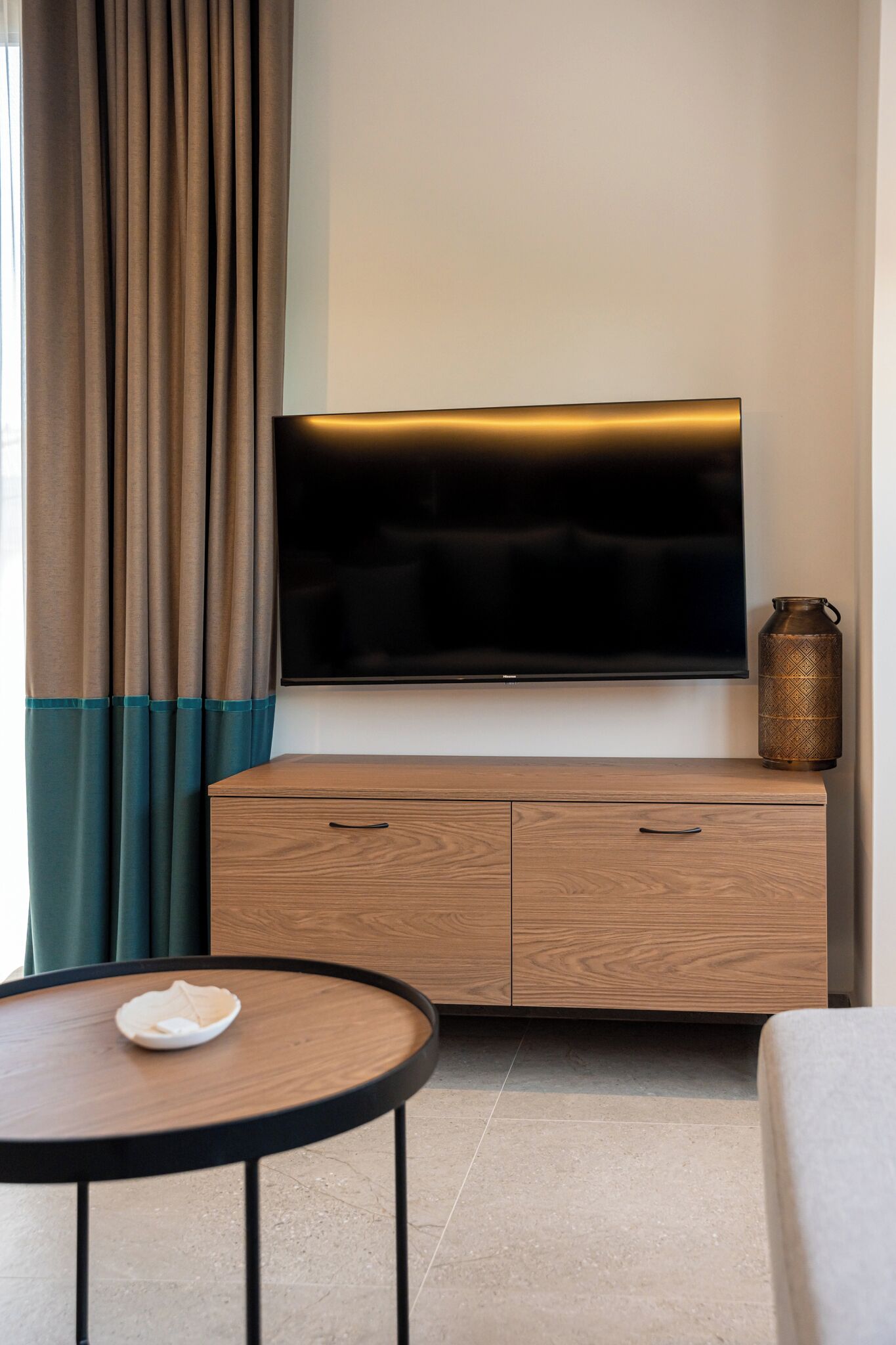 Ein modernes Hotelzimmer mit einem Flachbildfernseher an einem Holzschrank, einem runden Tisch mit weißem Teller und einer dekorativen Metallvase.