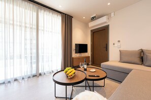 A modern hotel room with a sofa, a round table with a fruit bowl, a flat-screen TV, and a large window with sheer curtains. - Sea Breeze Luxury Apartments (Nea Propontida)