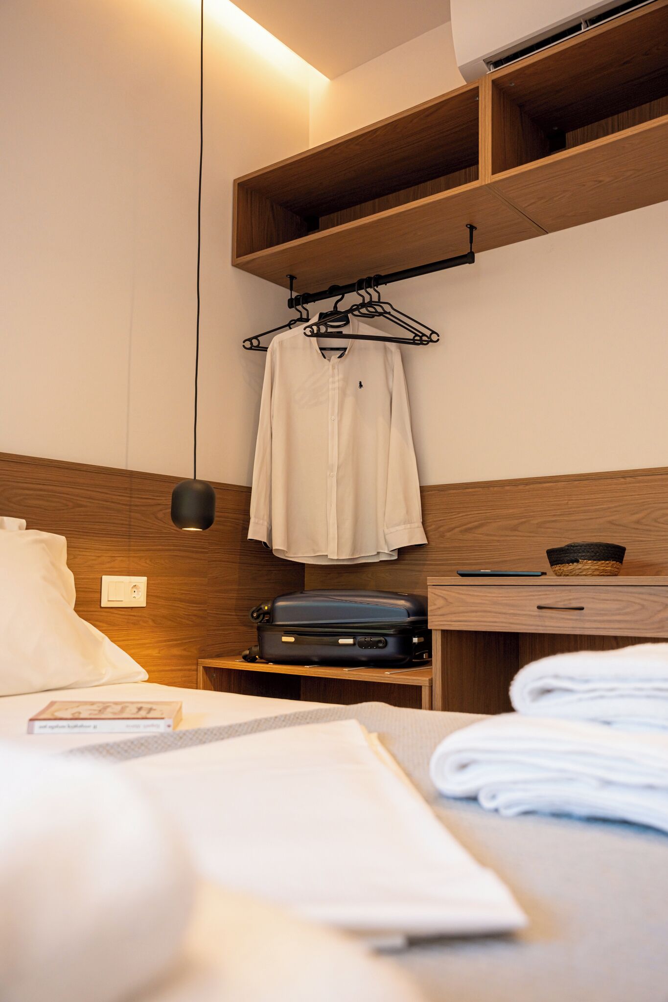 Ein Hotelzimmer mit einem Bett, einem Nachttisch mit Lampe, einem Koffer und einem Regal mit einem gefalteten Handtuch.