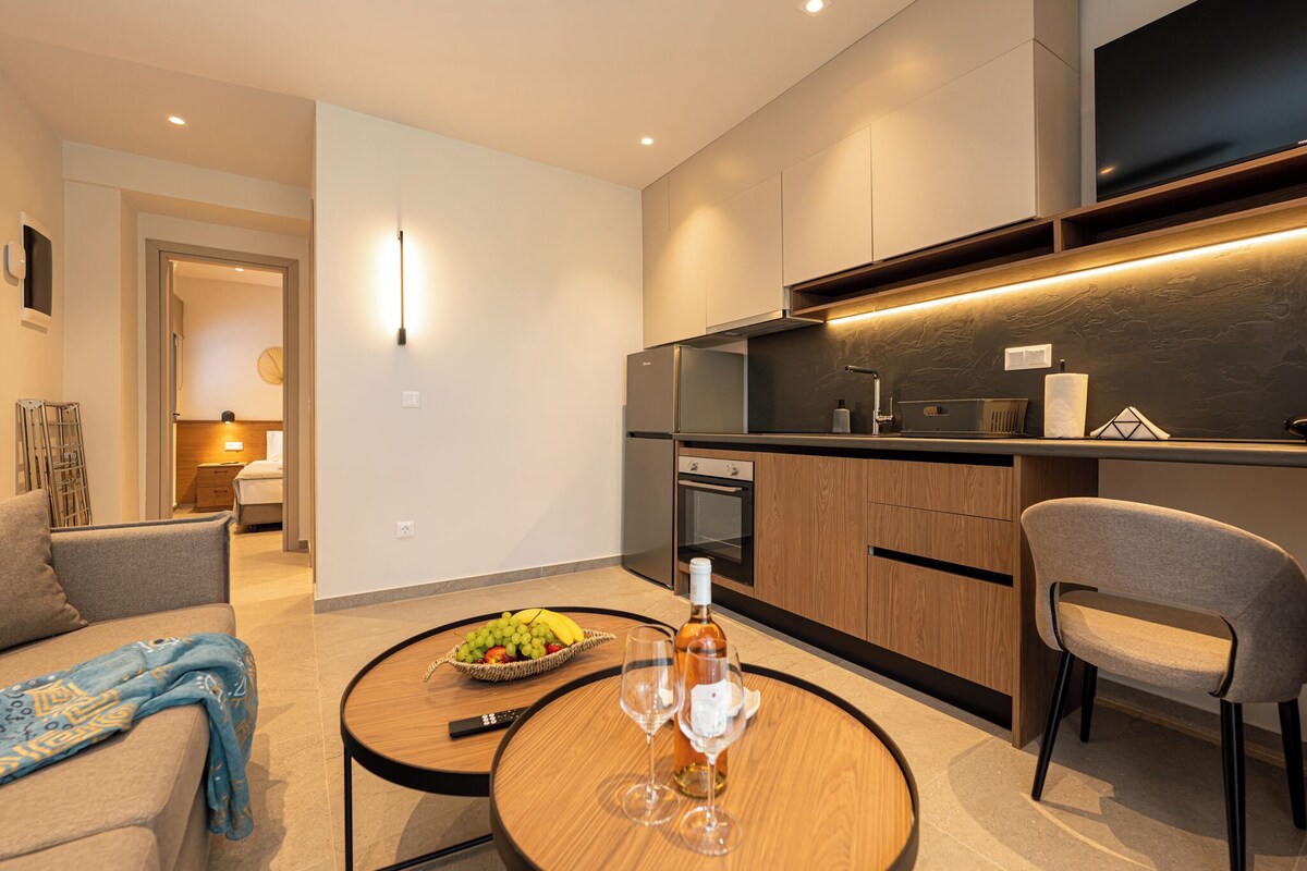 Una sala moderna con un sofá, una mesa de centro redonda con una botella y vasos, un comedor con una silla y una cocina con armarios de madera y un televisor.