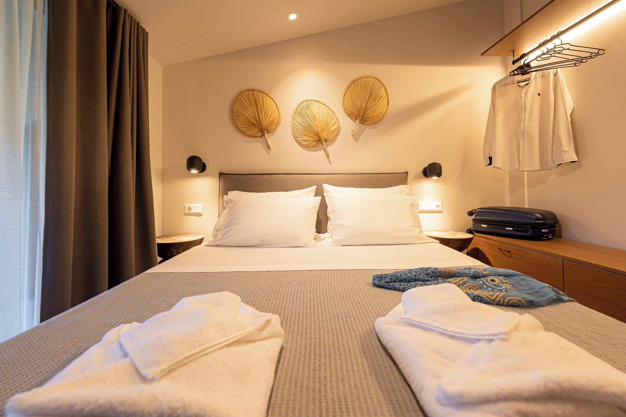 A hotel room with a bed, two bedside tables, a wall-mounted lamp, a suitcase, and a towel rack.