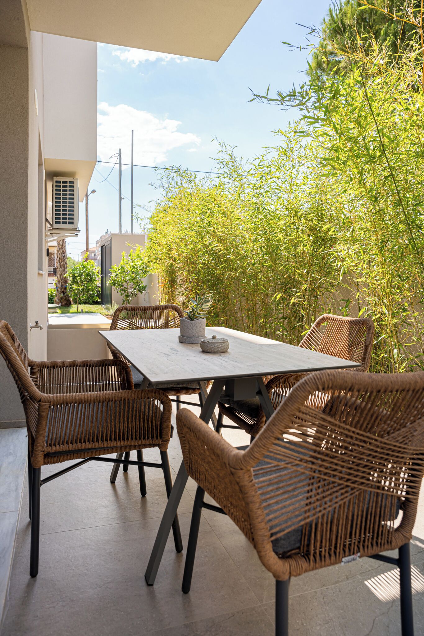 An outdoor patio with a table and wicker chairs, surrounded by greenery.
