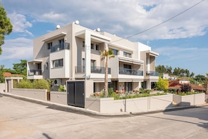 Un moderno edificio residencial de varios pisos con balcones, entrada porticada y un jardín ajardinado.
