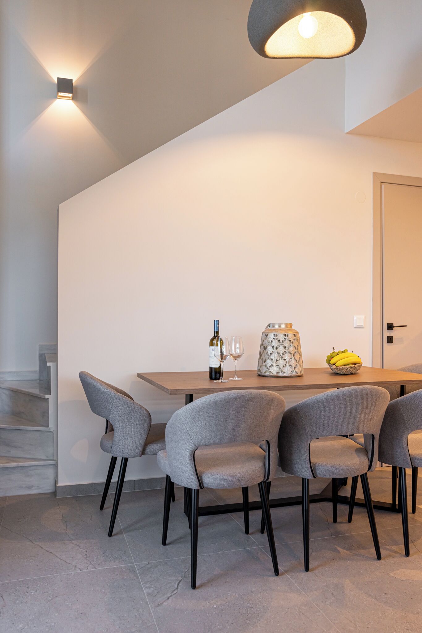 Un comedor moderno con una mesa de madera, sillas grises tapizadas, una botella de vino, dos copas, un jarrón decorativo y un cuenco de fruta.