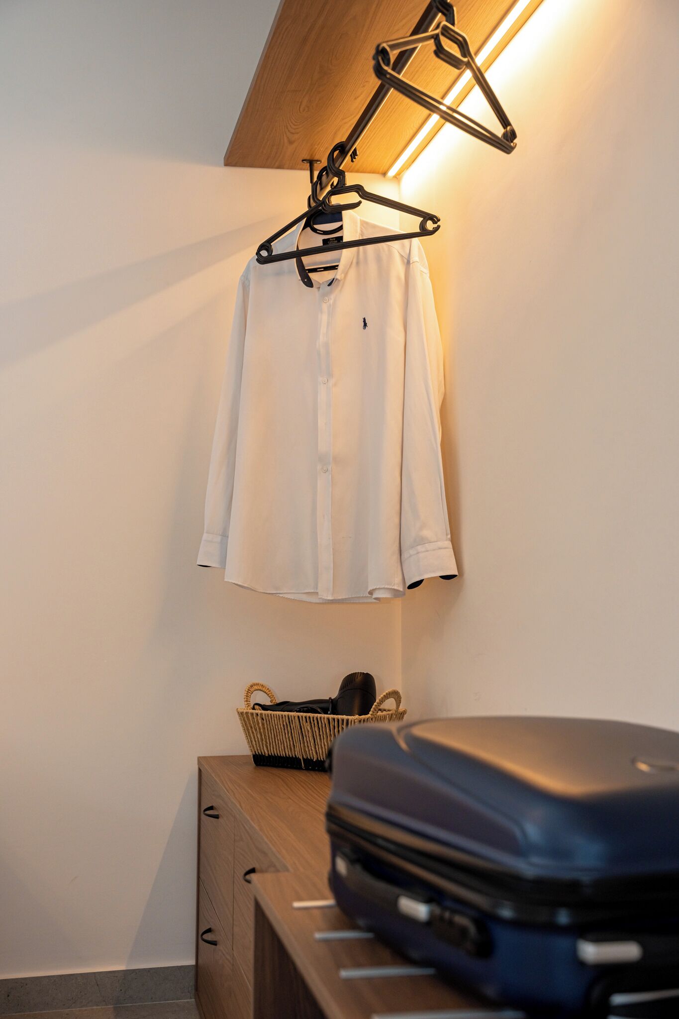 Uma camisa branca pendurada em um cabide preto num quarto com uma cômoda de madeira e um cesto.
