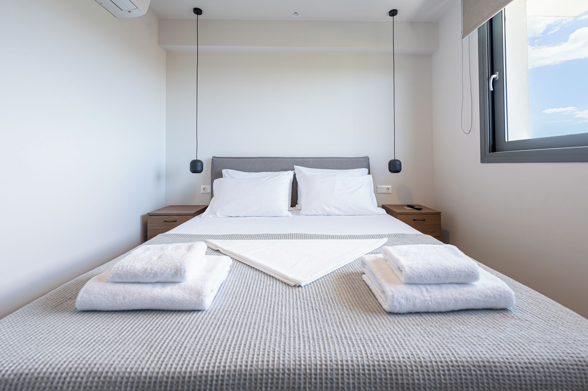 Een net opgemaakt bed met witte lakens, een grijs beddekleed met structuur en twee handdoeken gevouwen aan het voeteneind.
