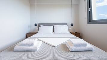 Een net opgemaakt bed met witte lakens, een grijs beddekleed met structuur en twee handdoeken gevouwen aan het voeteneind.