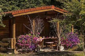 Eine rustikale Holzhütte mit einer überdachten Veranda, einem Tisch mit rot-weiß kariertem Tuch und leuchtend violetten Blumen in Pflanzgefäßen.