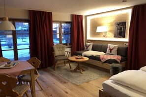 Ein gemütliches Zimmer mit Sofa, Ohrensessel, Esstisch und Blick auf eine verschneite Landschaft durch große Fenster.