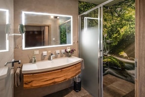 Ein modernes Badezimmer mit einem Holzkosmetikschrank, einem groĂen Spiegel mit Beleuchtung, einer Glasdusche und einem Wandbild mit Naturmotiv.