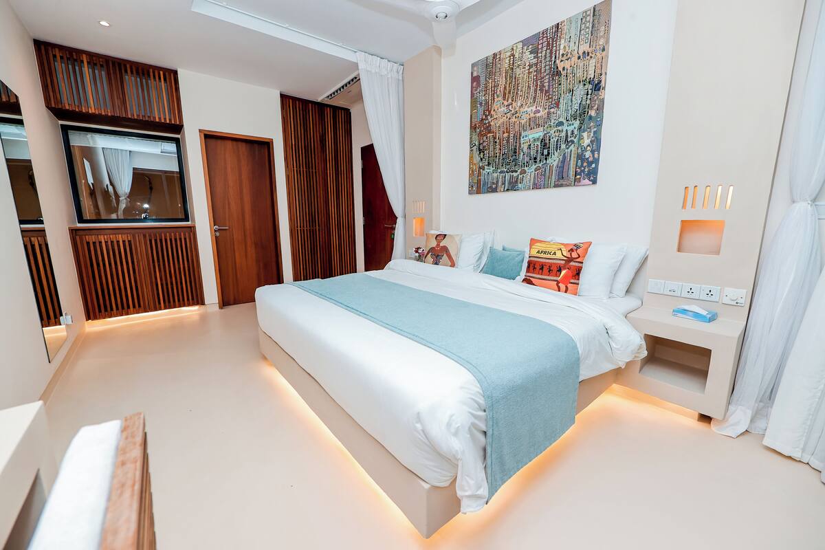 A modern bedroom with a large bed, a nightstand, a wall-mounted TV, and a window with curtains.