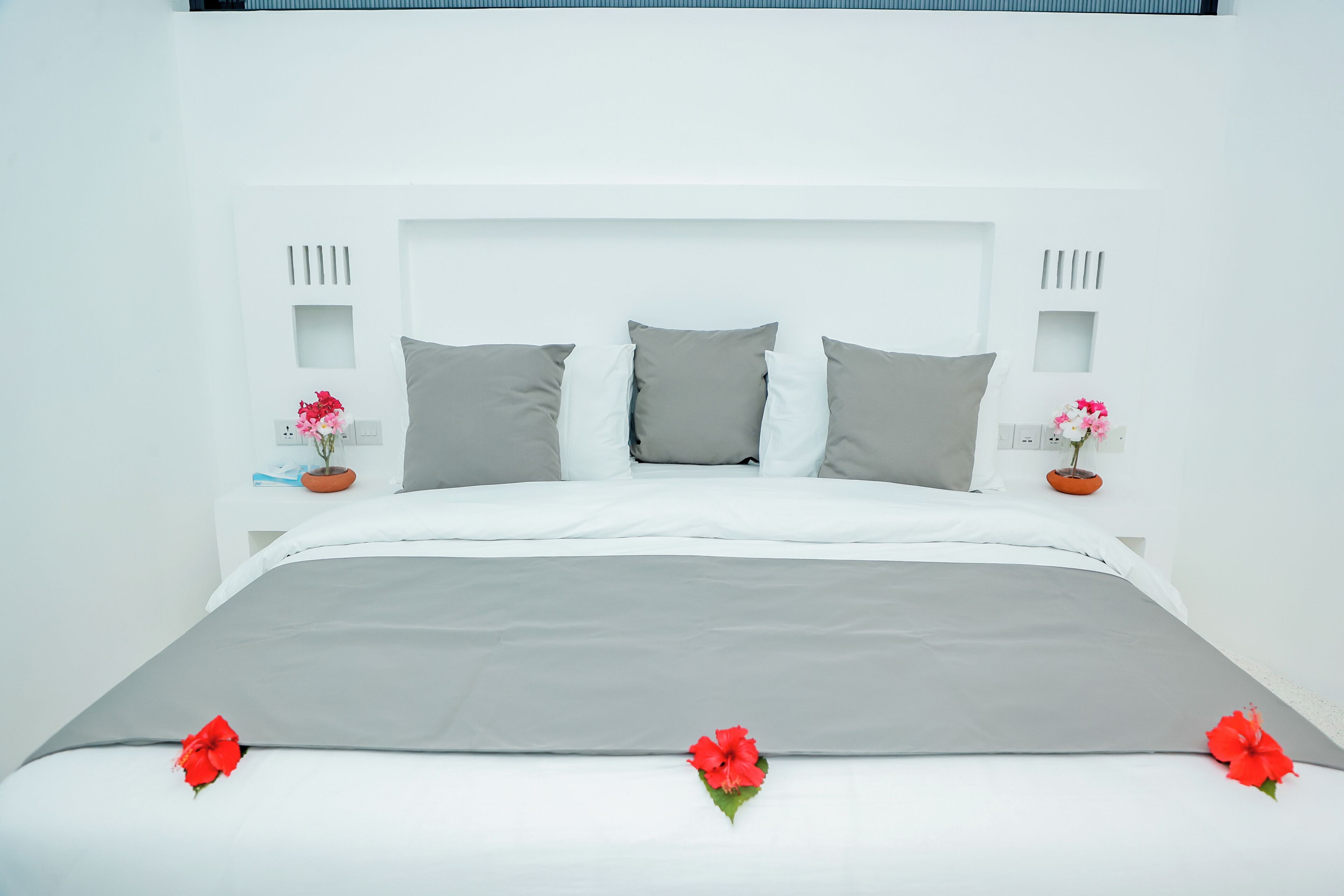 Una cama con un cubrecolcha gris, almohadas blancas y flores rojas.
