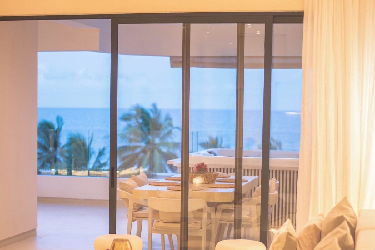 Habitación de hotel moderna con un gran ventanal corredero que da a un balcón con vista al océano y palmeras.