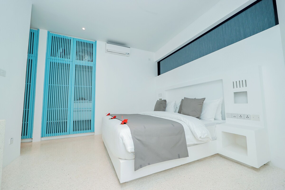 A modern bedroom with a white bed, grey bedding, and a blue window blind.