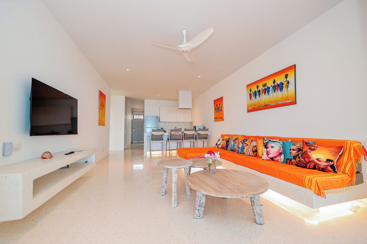 Una sala de estar moderna con un televisor de pantalla plana, un sofá blanco con cojines de colores, un ventilador de techo y una cocina al fondo.