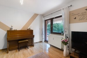 Ein Zimmer mit Klavier, Fernseher, Heizkörper und einem Fenster mit Vorhängen.