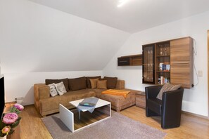 Ein Wohnzimmer mit einem Ecksofa, einem Couchtisch, einem schwarzen Sessel, einem Holzkabinett und einem Fernseher.