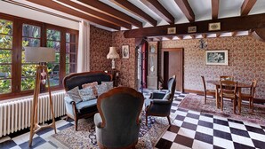 Een kamer met een geruite vloer, houten balken in het plafond, een radiator, een bank, een stoel, een tafel met stoelen en een raam met uitzicht op groen.