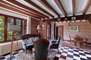 A room with a checkered floor, wooden ceiling beams, a radiator, a sofa, a chair, a table with chairs, and a window with a view of greenery.