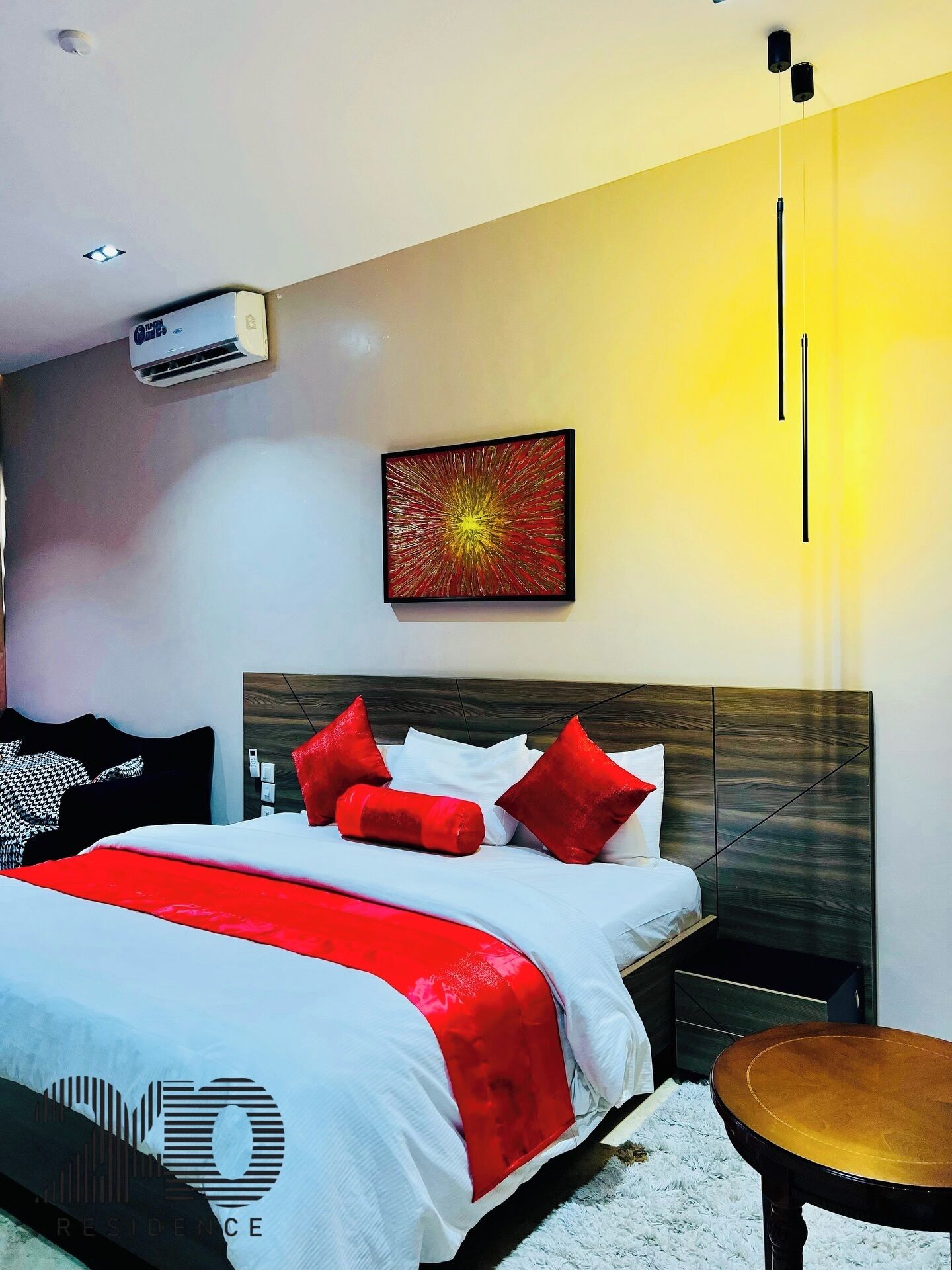 A modern hotel room with a large bed, a bedside table, a wall-mounted air conditioner, and a painting.