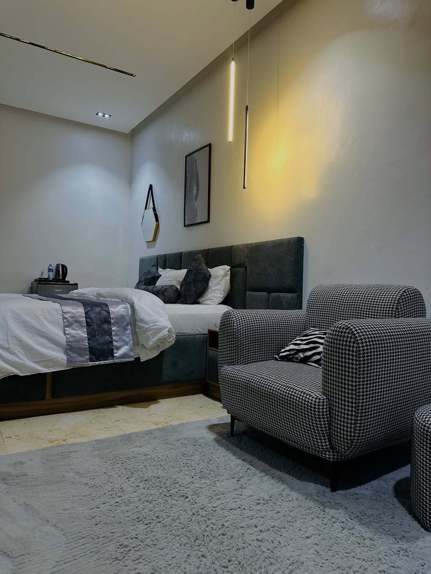 A modern bedroom with a bed, a houndstooth patterned armchair, a bedside table with a kettle, and a wall-mounted light fixture.