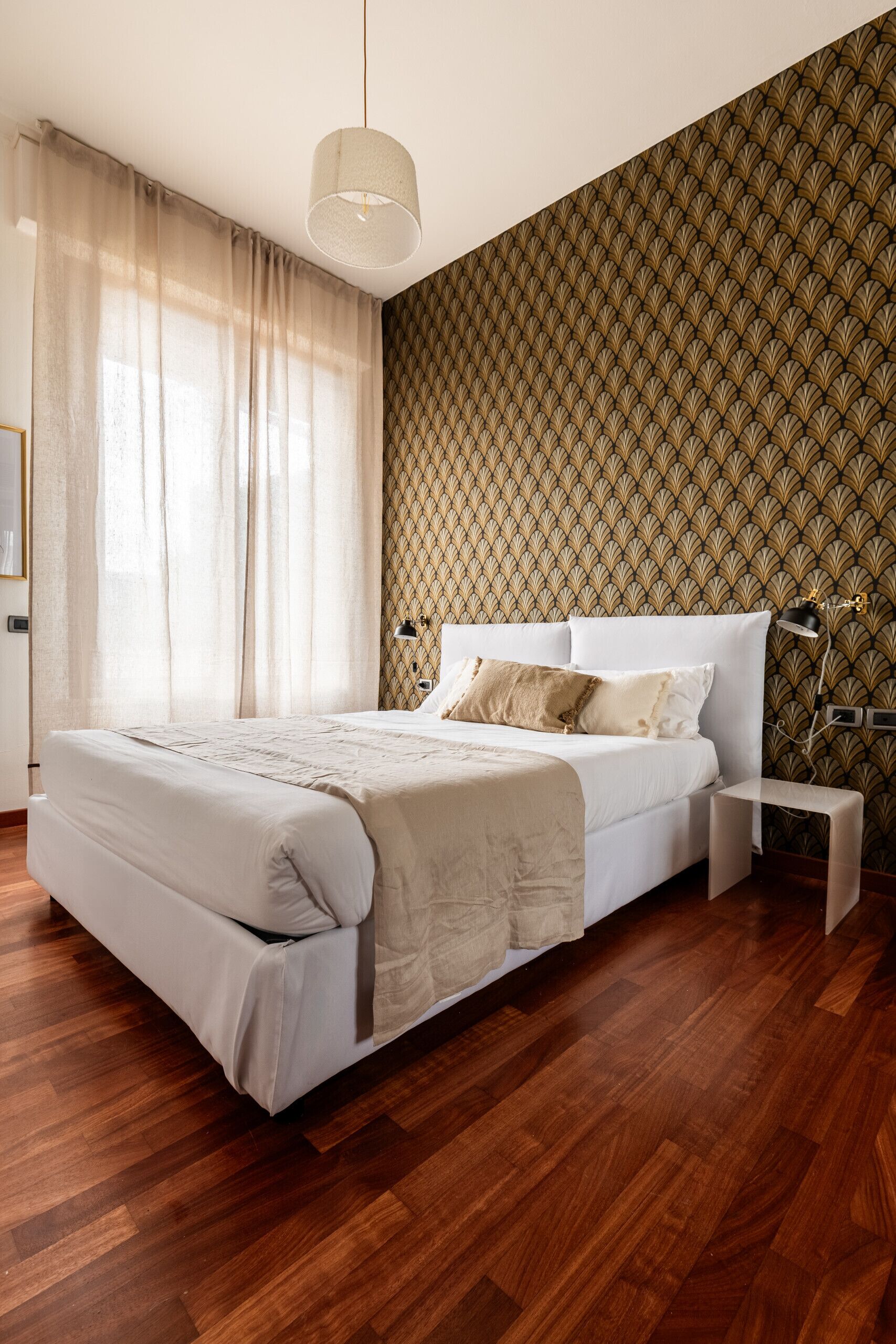 Ein Schlafzimmer mit Holzboden, einem Bett mit weißer Bettwäsche, einem Nachttisch, einer Lampe und einem Wandpapier mit Muster.