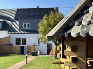 Een houten hutje met een schuin dak, een heldere lucht en een huis met een donker dak op de achtergrond.