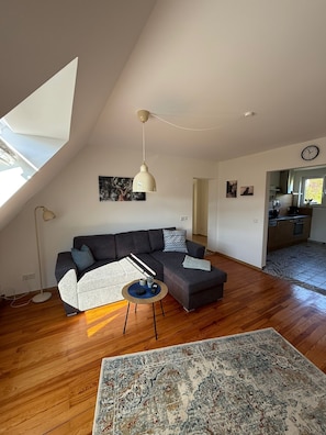 Een modern loft appartement met een grote L-vormige hoekbank, een ronde salontafel, een kamerkleed met een patroon, een hanglamp en een keuken op de achtergrond.