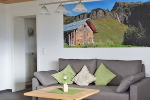 Ein graues Sofa mit grünen und weißen Kissen, ein kleiner Holztisch mit einer Vase und ein Berglandschaftsbild darüber.