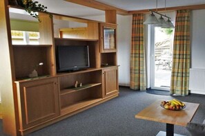 Ein Wohnzimmer mit Fernseher, Holzregalen, einem Tisch mit Obst und karierten Vorhängen.