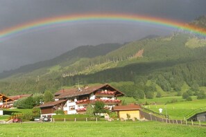Un chalet doté d’un balcon, entouré de collines verdoyantes et où l’on peut admirer un arc-en-ciel.