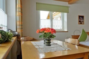 Ein Zimmer mit Holztisch, Blumen in einer Vase, einem Fenster mit Aussicht und einem Bett mit Kissen.