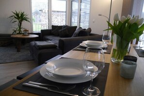 Ein Essbereich mit Tisch für zwei Personen, einer Blumenvase und Blick ins Freie durch große Fenster.