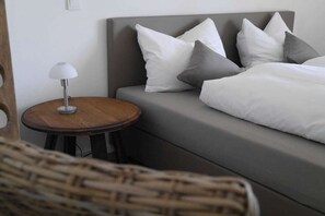 Ein Bett mit weißen und grauen Kissen, ein runder Holztisch mit einer Lampe sowie ein Korbstuhl.