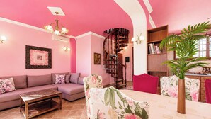 Un salon décoré dans des tons roses, avec un canapé, une table basse, des fauteuils à motifs floraux, un escalier et une bibliothèque.