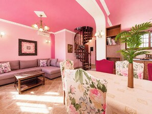 A pink-themed living room with a sofa, coffee table, floral chairs, a staircase, and a bookshelf.