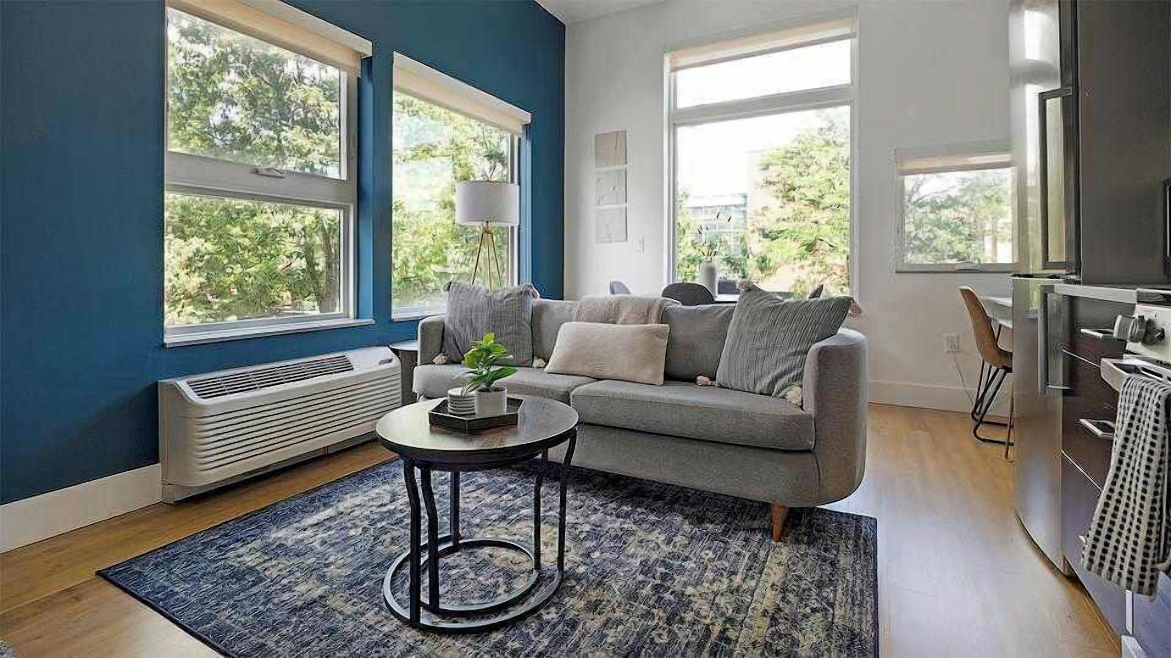 A living room with a grey sofa, a round coffee table, and a blue rug. There are large windows with blinds, a white radiator, and a kitchen area with a stove.
