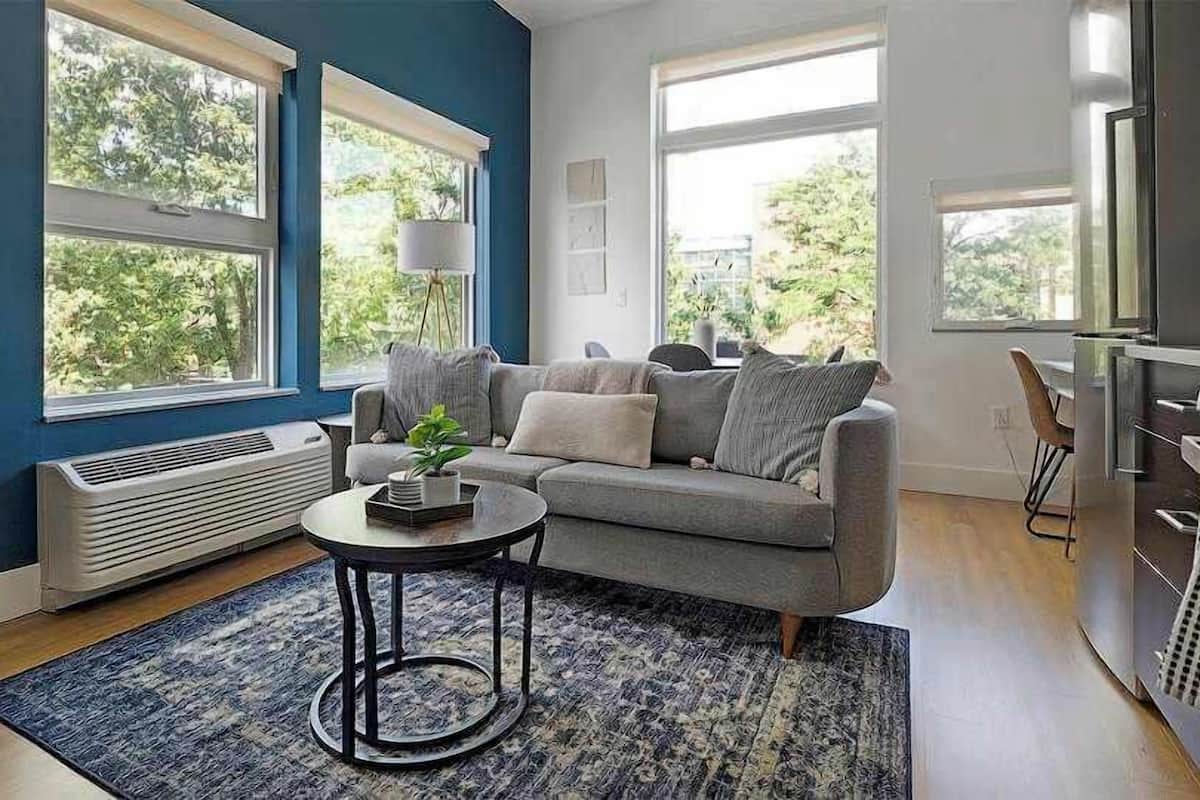 A living room with a grey sofa, a round coffee table, and a blue rug. There are large windows with blinds, a white radiator, and a kitchen area with a stove.