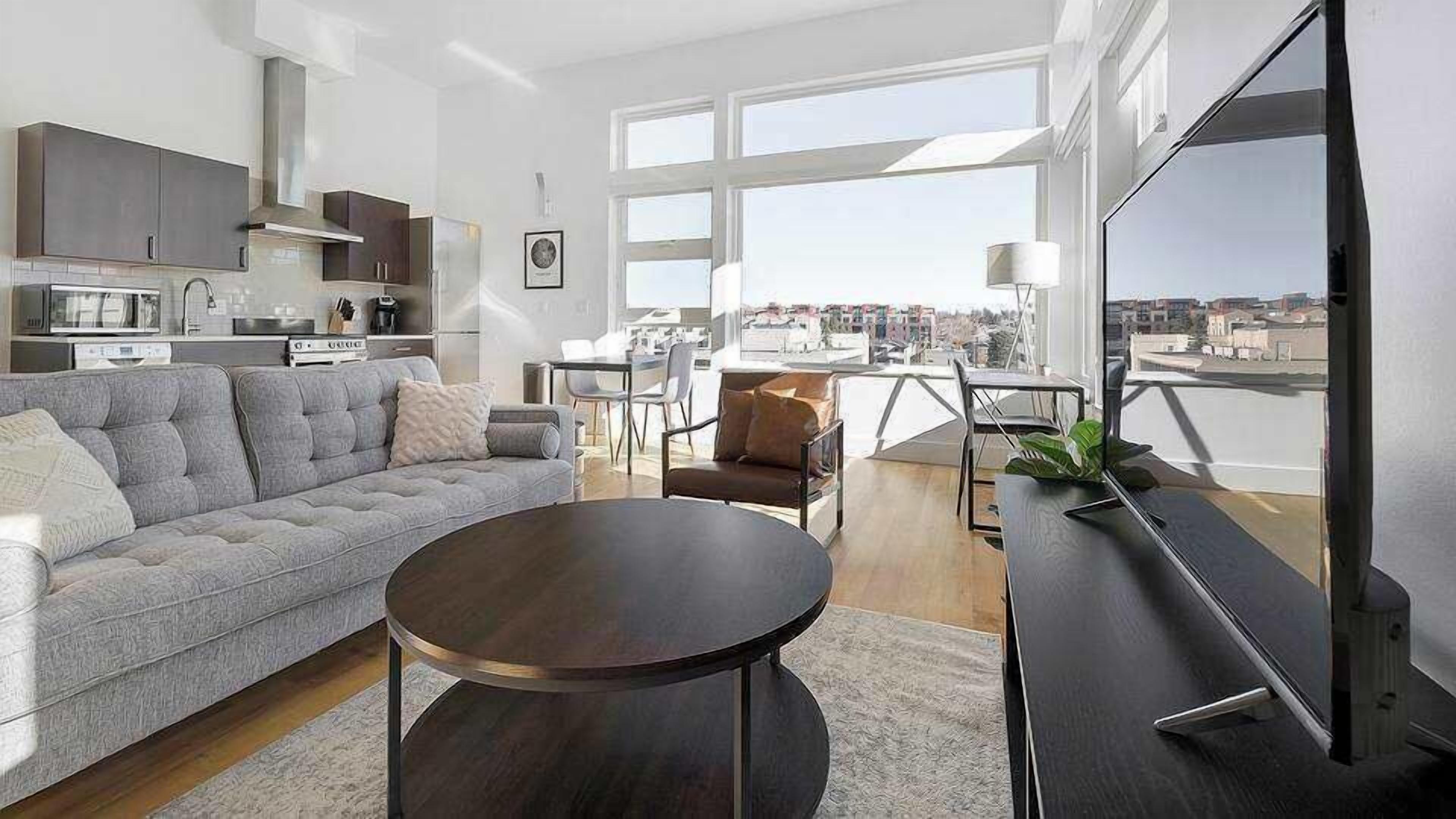 A modern living room with a sofa, coffee table, and TV. There is a kitchen area with cabinets and appliances. The room has large windows offering a city view.