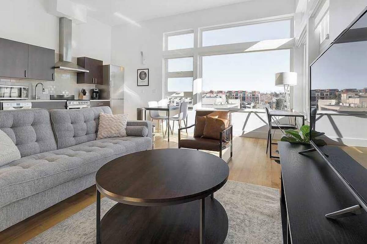 A modern living room with a sofa, coffee table, and TV. There is a kitchen area with cabinets and appliances. The room has large windows offering a city view.
