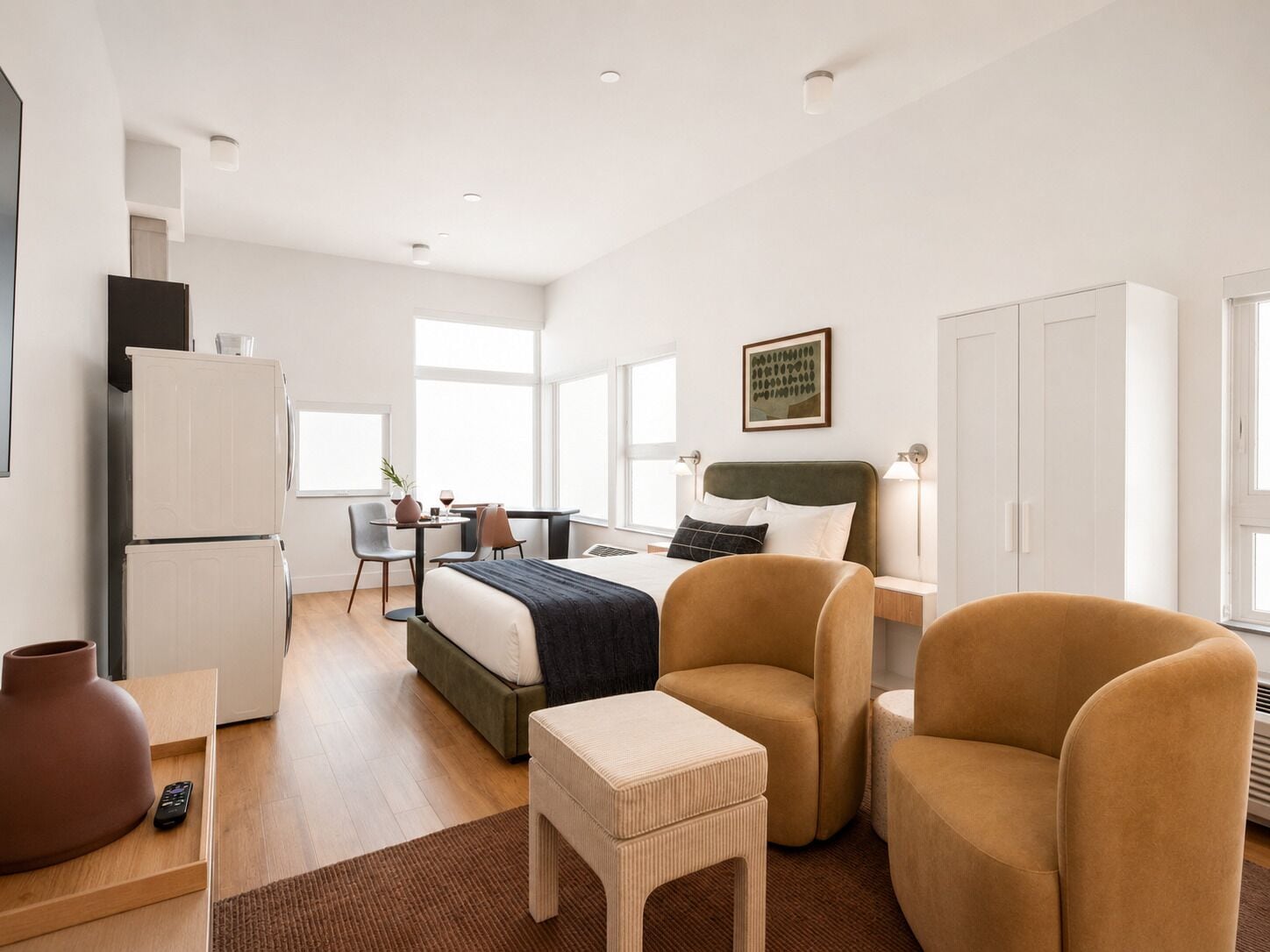 Una habitación de hotel moderna con una cama, dos sillones, una mesita, un refrigerador y un ventanal que proporciona luz natural.
