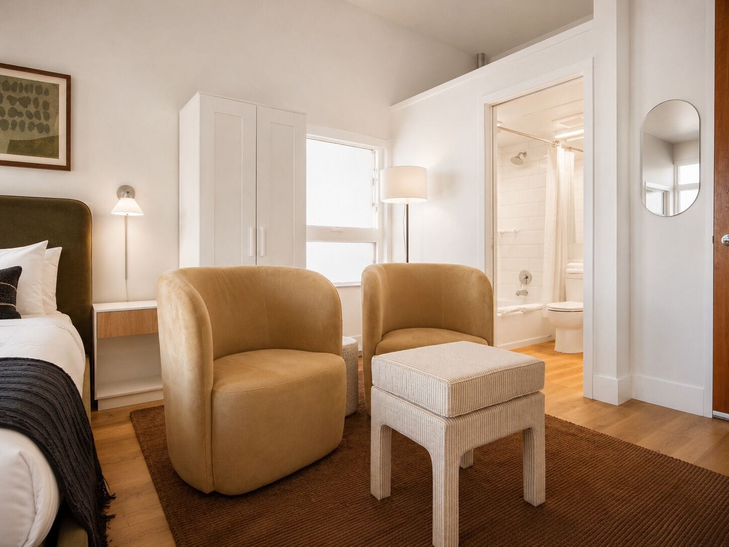Habitación de hotel con cama, dos sillones, una mesita, lámpara, espejo y baño visible a través de una puerta abierta.