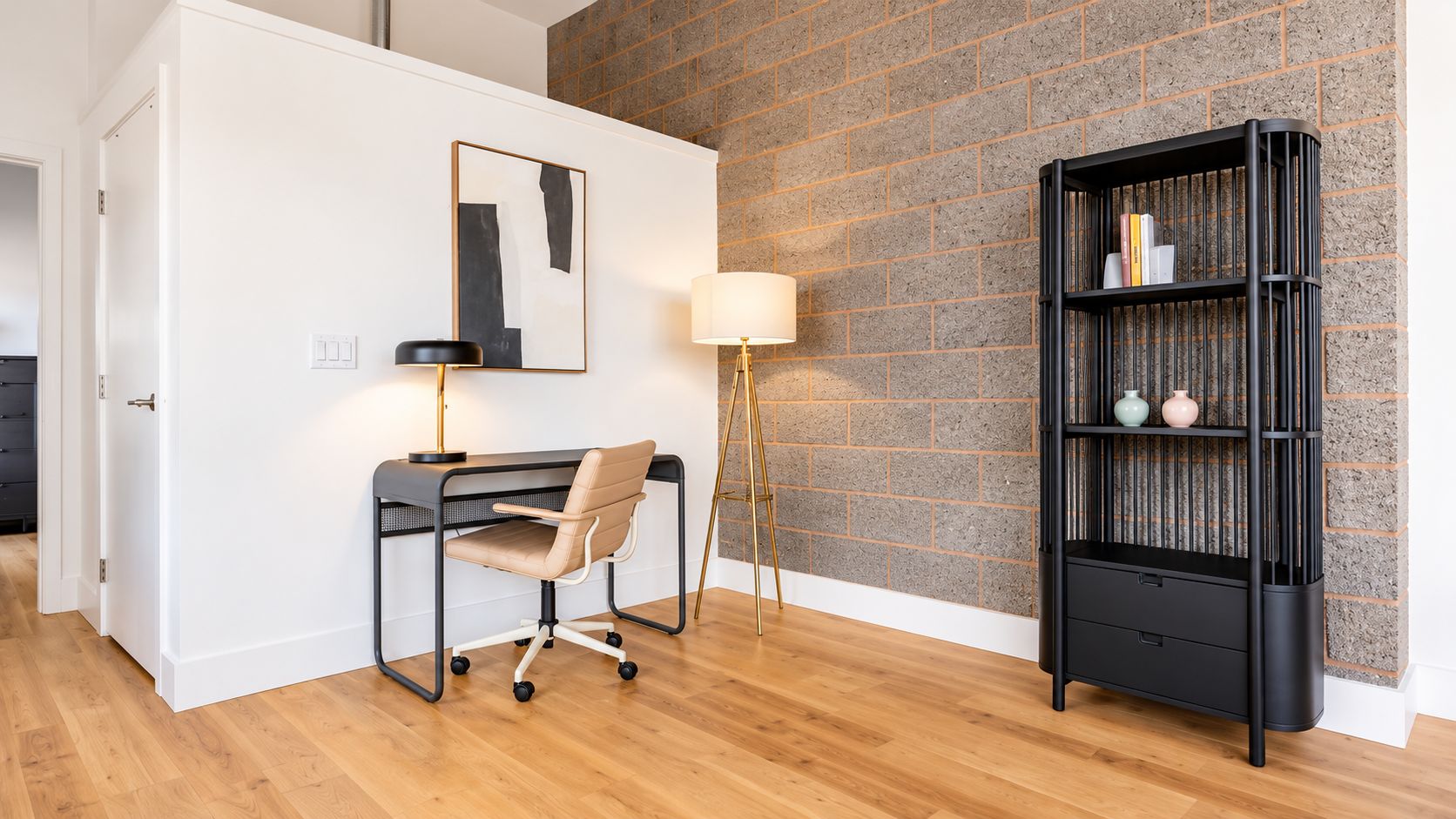 A room with a desk, chair, lamp, and bookshelf against a brick wall.